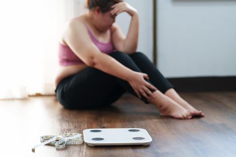 Kränkung durch Übergewicht: Frau sitzt traurig hinter Waage