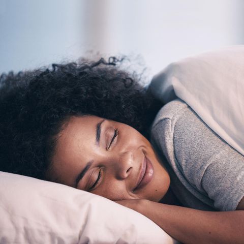 Frau mit Afro liegt glücklich und zufrieden im Bett und schläft. Ihre Lippen sind zu einem zufriedenen Lächeln geformt.