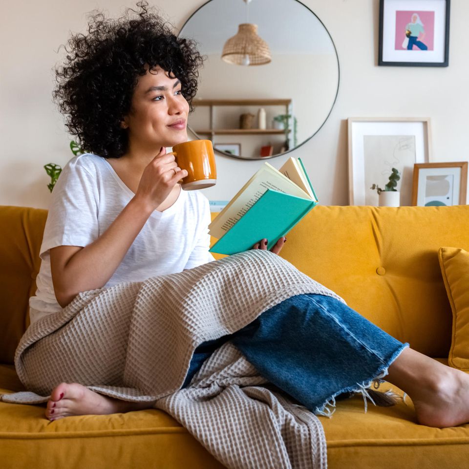 Eine Frau sitzt mit Buch und Tasse auf dem Sofa