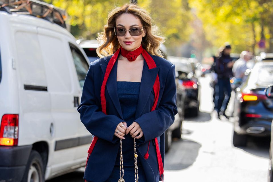 Auch in diesem Herbst lassen die Streetstyles anlässlich der Paris Fashion Week nichts zu wünschen übrig. Außerhalb der Miu Miu Show sehen wir Merve Görgöz in einer Kombination in Navy-Blau. Sie besteht aus einem langen Blazer und einem Bandeau-Maxikleid. Einen Kontrast bieten rote Farbakzente von Schal und Schuhen, welche jeweils mit angenähten Stoffrosen überzeugen. Als Designer-Pieces trägt sie eine große Brille von Miu Miu und eine metallische Tasche von Paco Rabanne.