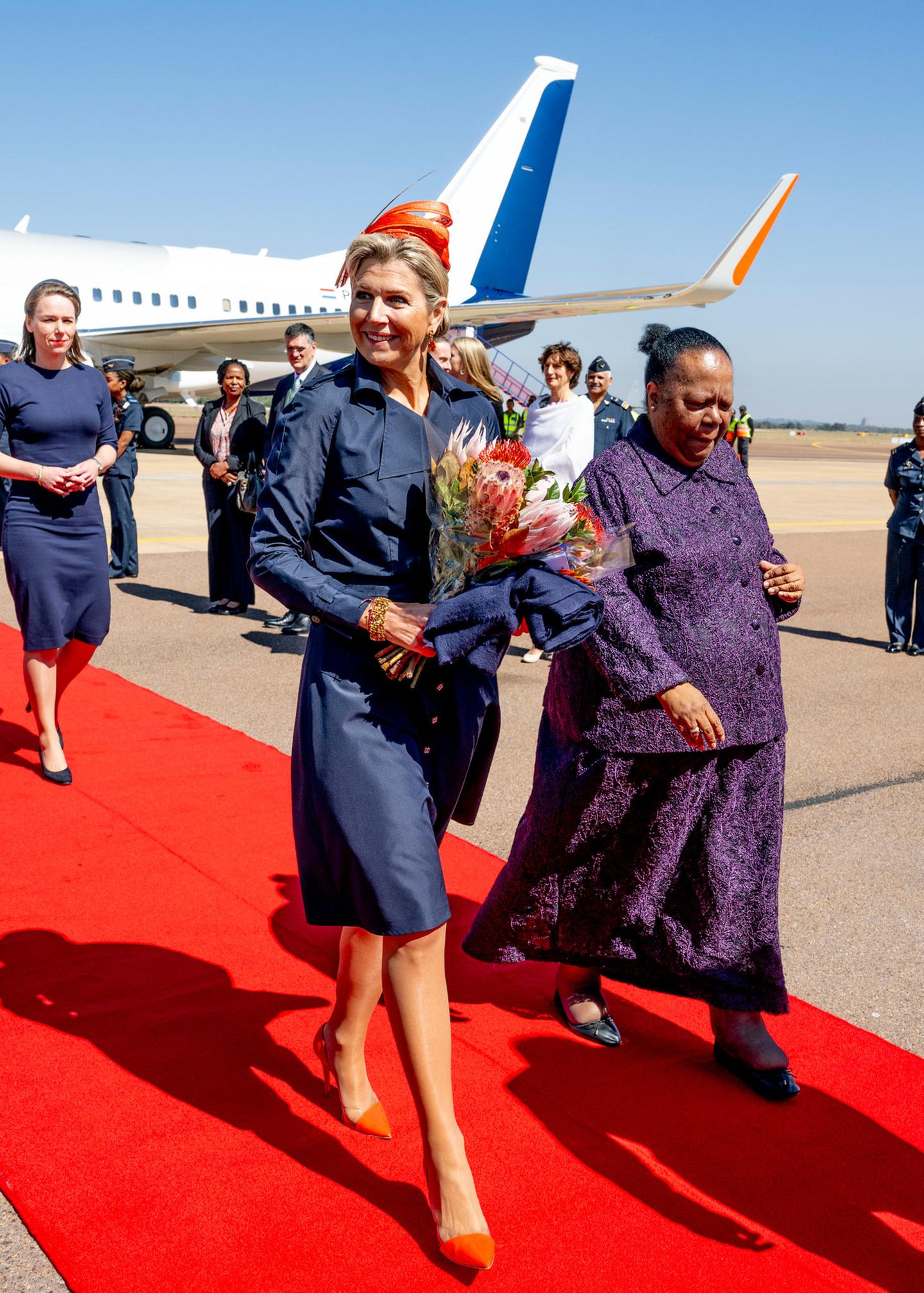 Star-Auftritt: Königin Máxima besucht erstmals im offiziellen Rahmen Südafrika und begeistert bei ihrer Ankunft in Pretoria in einem stilvollen Mantelkleid, das sie mit einem leuchtend orangefarbenen Kopfschmuck kombiniert. Dazu stylt sie passende semitransparent-orangene Pumps von Maison Natan Couture. Doch bei denen soll es nicht blieben. 