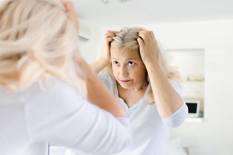 Haarausfall und Wechseljahre: Eine mittelalte Frau schaut im Spiegel skeptisch auf ihren Scheitel