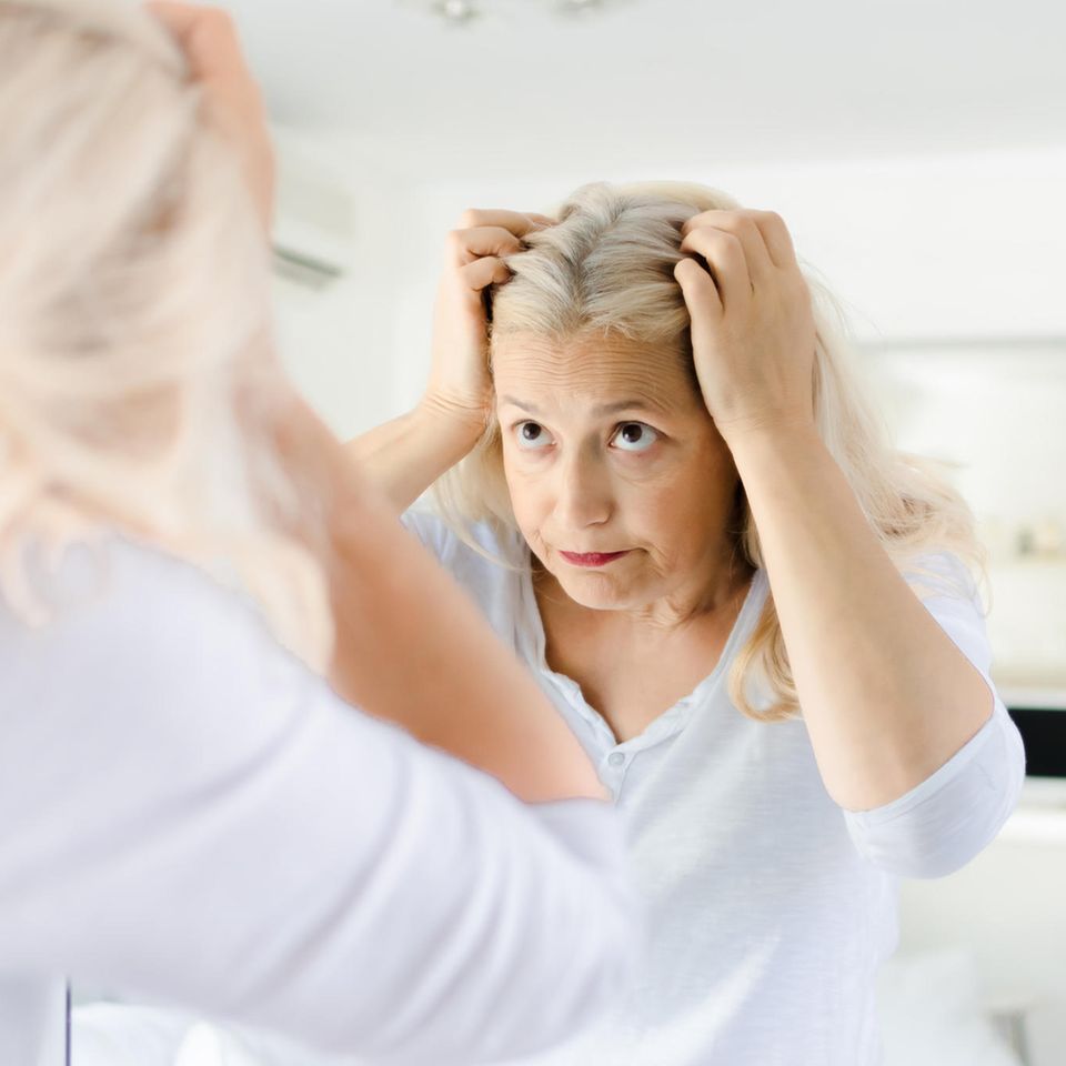 Haarausfall und Wechseljahre: Eine mittelalte Frau schaut im Spiegel skeptisch auf ihren Scheitel