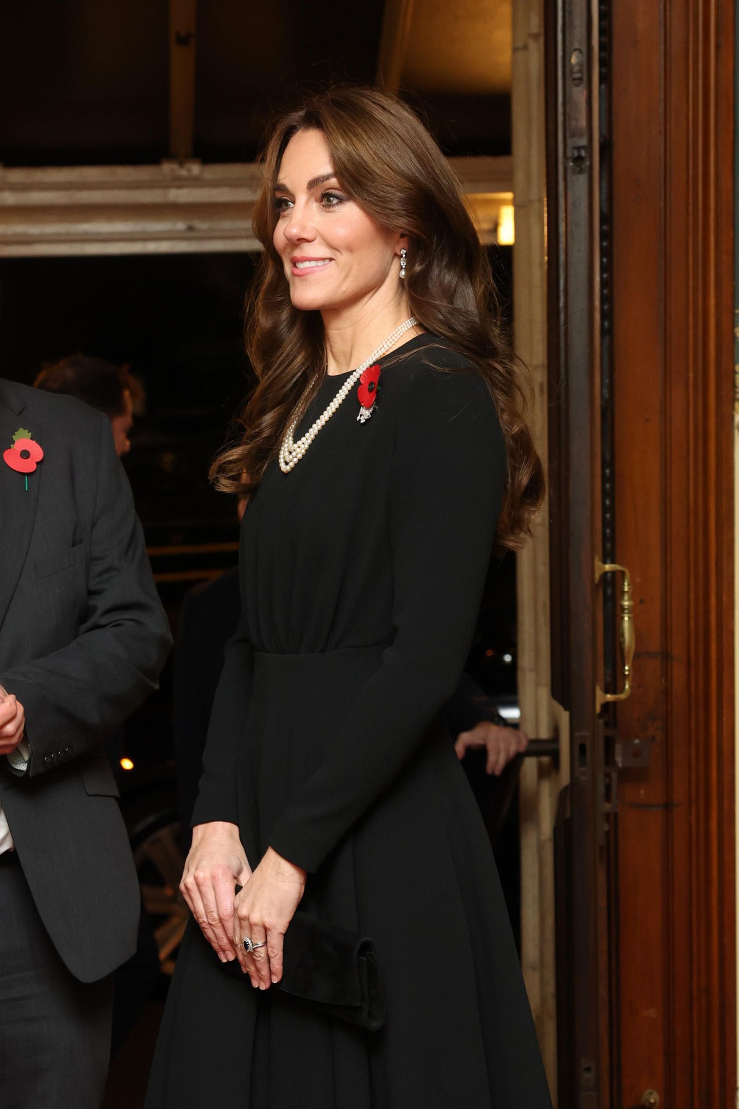 Das schwarze Kleid von Emilia Wickstead ist ebenso schlicht. Die edle Perlenkette von Queen Elizabeth fügt sich hervorragend in den Look, der mit Eleganz besticht. Natürlich trägt Kate auch hier einen Mohnblumen-Stecker. Ihre Haare hat sie in mühelose Wellen gestylt – die Karamell-Note in ihrer Haarfarbe unterstreicht die warme Ausstrahlung der Princess of Wales. 
