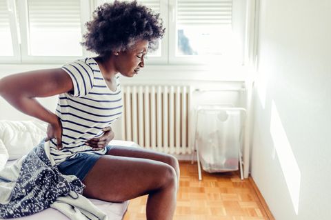 Unterleibsschmerzen in Wechseljahren: Schwarze Frau sitzt auf der Bettkante, hält sich vor Schmerzen Bauch und Rücken