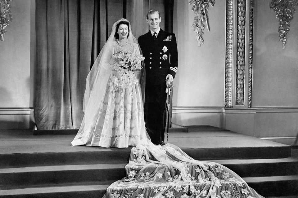 Am 20. November 1947 treten Queen Elizabeth und Prinz Philip vor den Traualtar. Norman Hartnell entwarf für die im September 2022 verstorbene Queen ein Kleid aus weißem Satin mit einem hohen Kragen und einer langen, mit Blumen bestickter Schleppe, die für die offiziellen Fotos dramatisch auf den Stufen vorm Brautpaar drapiert wurde. 
