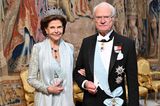 Königin Silvia hat sich farblich der Schärpe ihres Mannes, König Carl Gustafs, angepasst. Sie setzt auf ein mit Pailletten besticktes Kleid, zu dem sie eine Stola sowie eine Clutch in Muschelform von Judith Leiber im selben Farbschema wählt. 