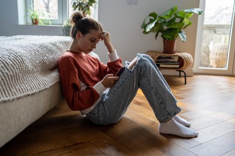 Einsamkeit Symptome: junge Frau sitzt traurig vor dem Bett und starrt aufs Handy