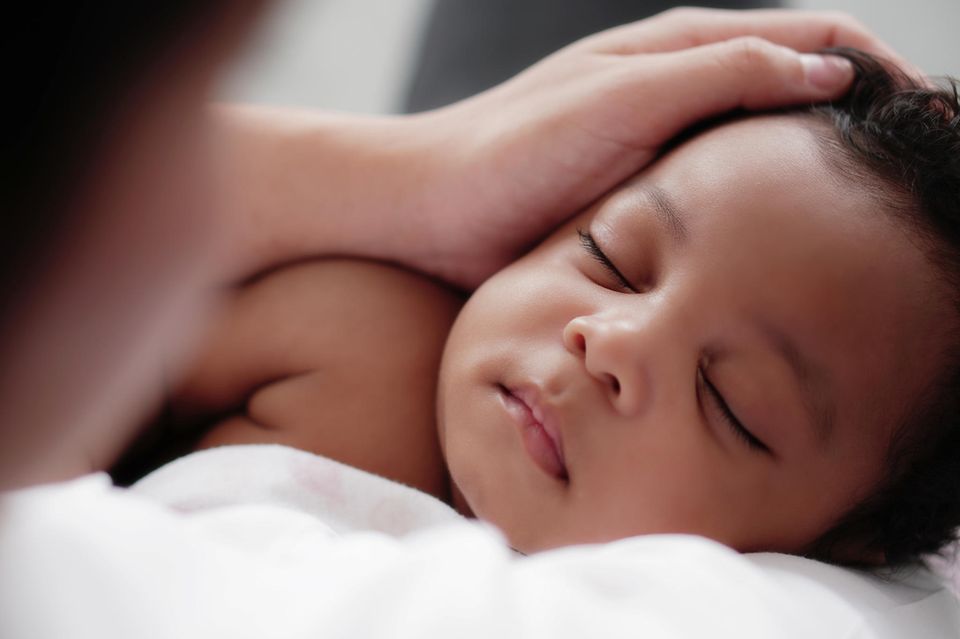 Stiko-Empfehlung für Meningokokken B: Ein schlafendes dunkelhäutiges Baby, eine Hand streichelt vorsichtig seinen Kopf
