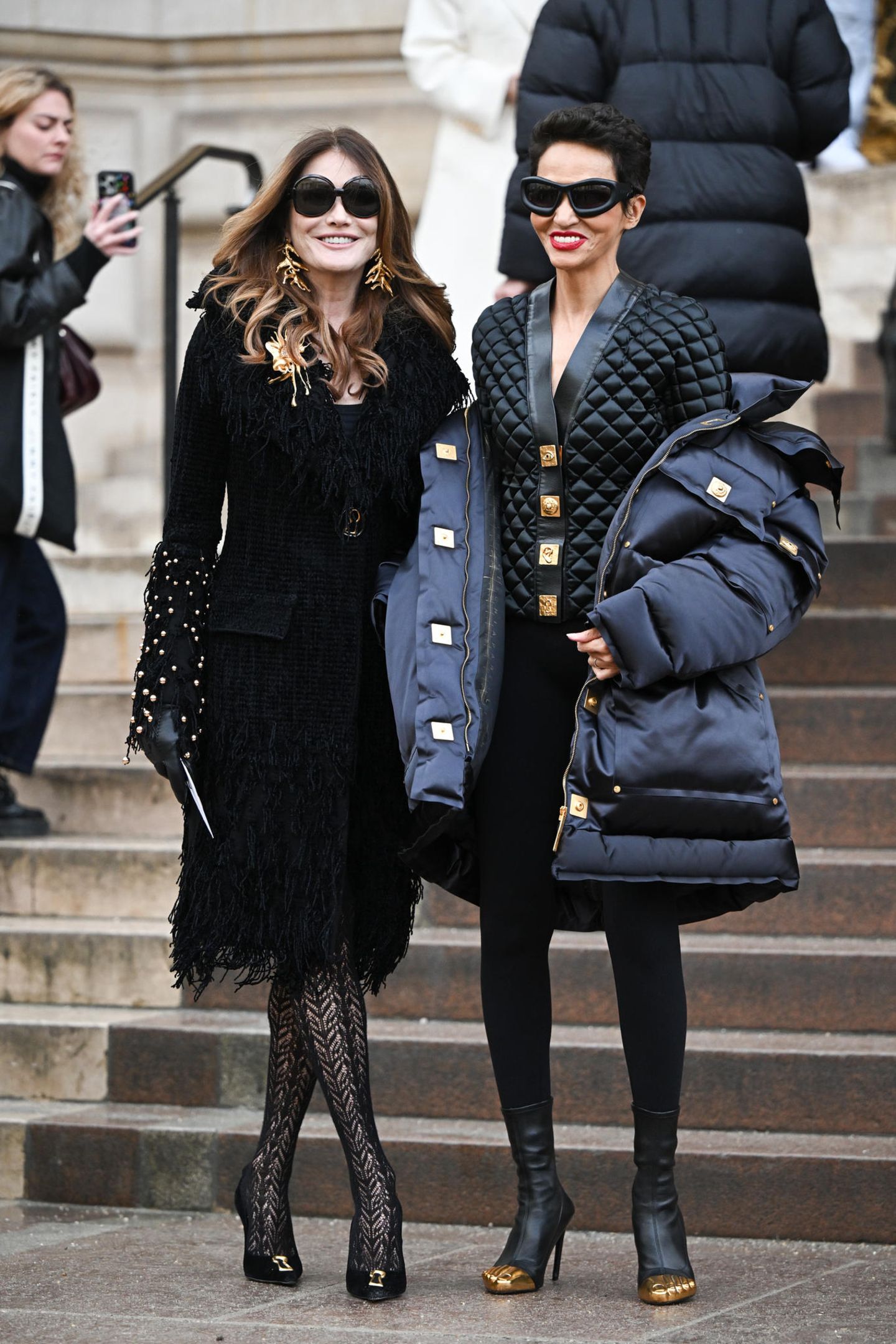 Carla Bruni und Farida Khelfa beweisen mit ihren schwarzen High-Fashion-Looks ihr Können als Fashionistas. Während Carla auf Korallen-Schmuck, Netzstrumpfhose und Fransenmantel setzt, hebt sich Farida mit dickem Outdoor-Mantel und Stepp-Blouson gekonnt von der Masse ab.