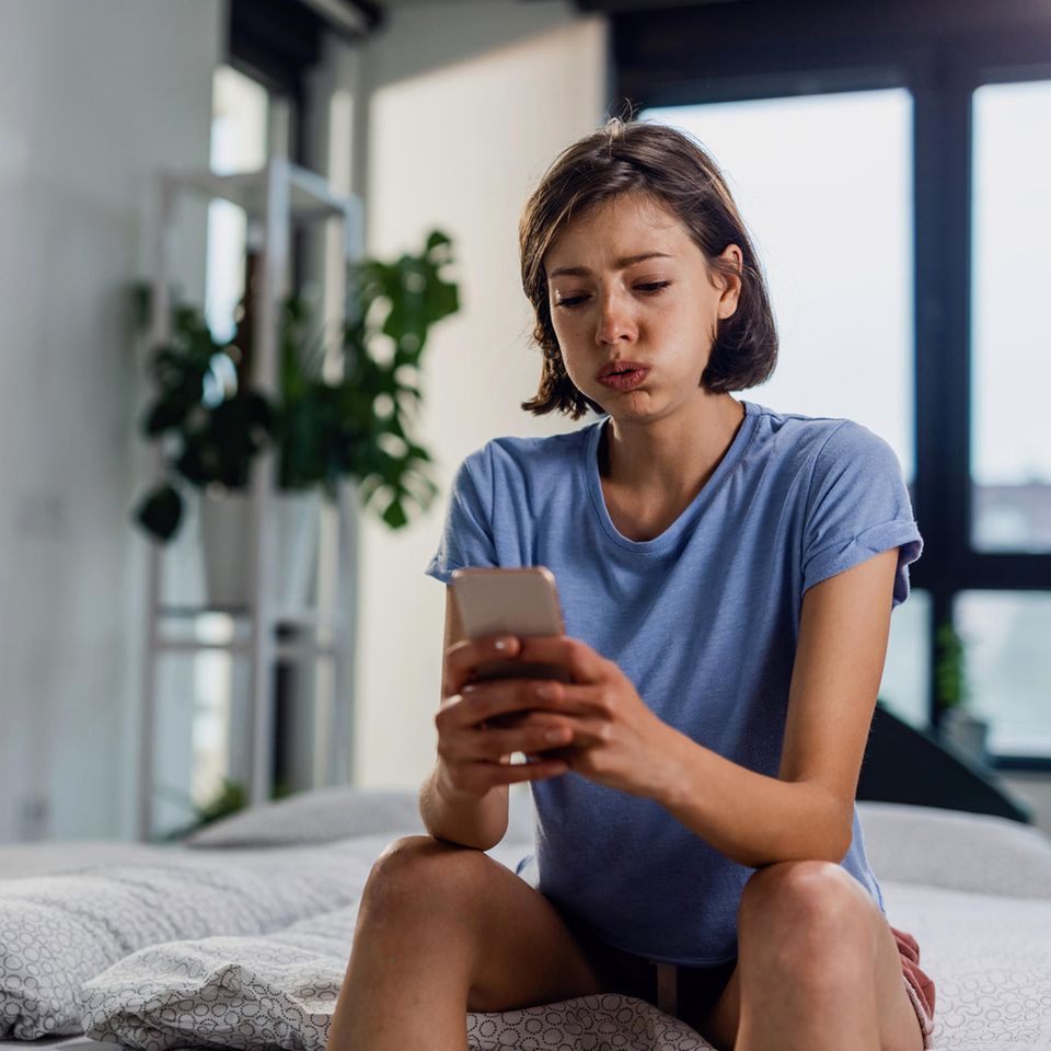 Dating-Apps: junge Frau sitzt frustriert mit dem Handy auf dem Bett