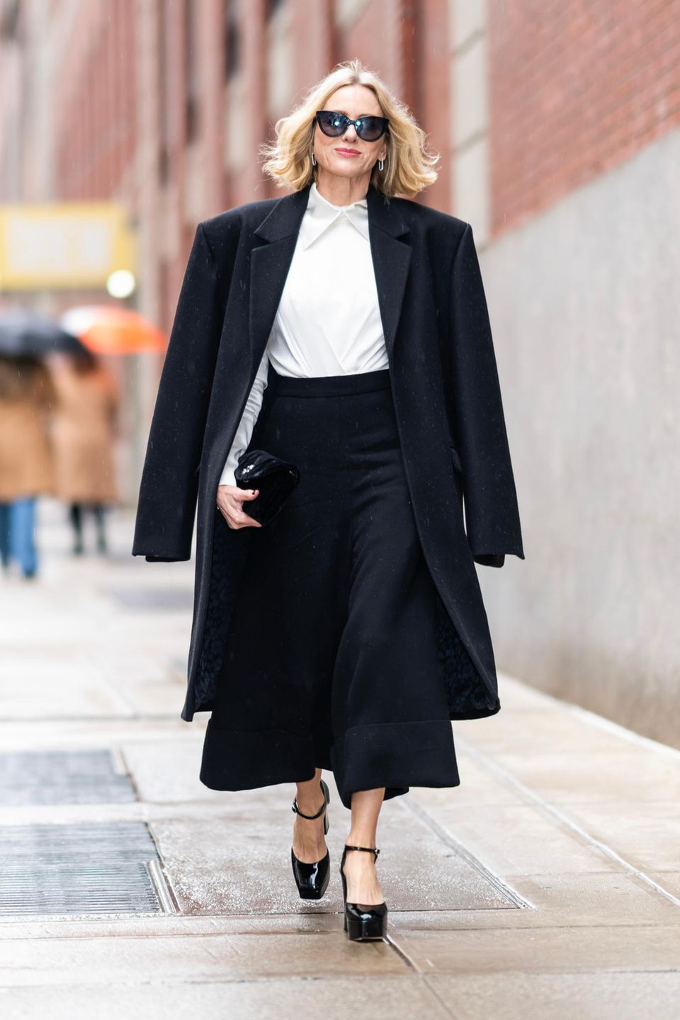 Pure Eleganz! In diesem klassischen Schwarz-Weiß-Look stolziert Schauspielerin Naomi Watts durch die Straßen von New York. Sie trägt eine schwarze Culotte-Hose, eine weiße Bluse und glänzende Plateau-Pumps. Stilbewusst wirft sie ihren schwarzen Mantel über ihre Schultern und rundet ihr Outfit mit einer großen Sonnenbrille sowie silbernen Ohrringen ab. Klassisch, elegant und ultra cool: Naomi trägt den Go-To-Look fürs Büro.
