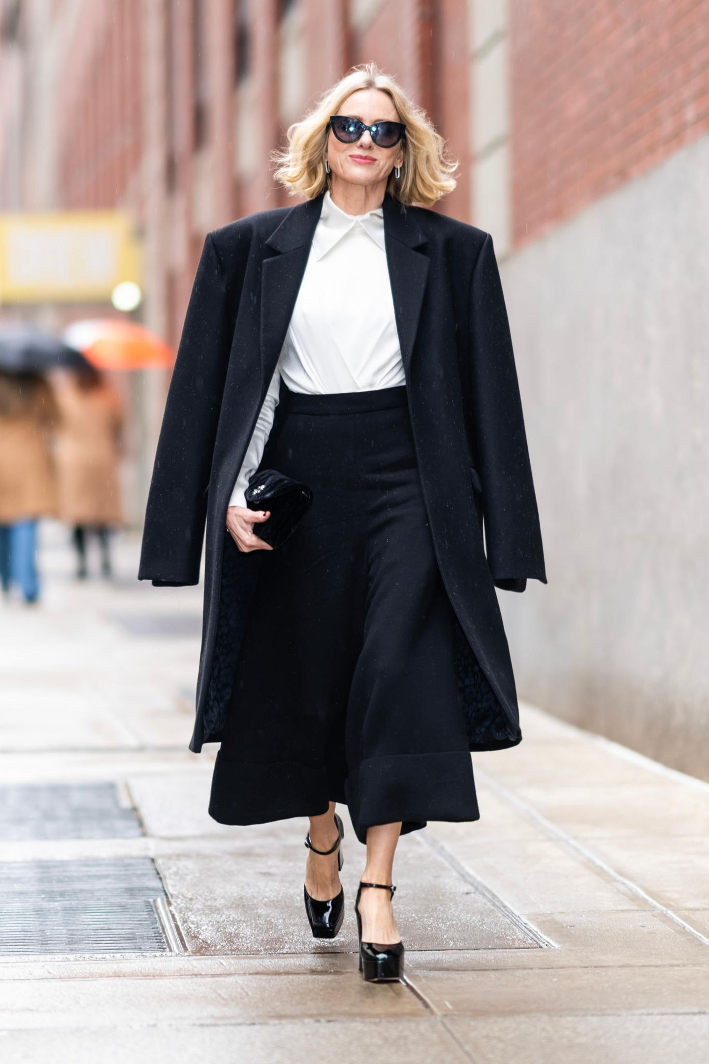 Pure Eleganz! In diesem klassischen Schwarz-Weiß-Look stolziert Schauspielerin Naomi Watts durch die Straßen von New York. Sie trägt eine schwarze Culotte-Hose, eine weiße Bluse und glänzende Plateau-Pumps. Stilbewusst wirft sie ihren schwarzen Mantel über ihre Schultern und rundet ihr Outfit mit einer großen Sonnenbrille sowie silbernen Ohrringen ab. Klassisch, elegant und ultra cool: Naomi trägt den Go-To-Look fürs Büro.