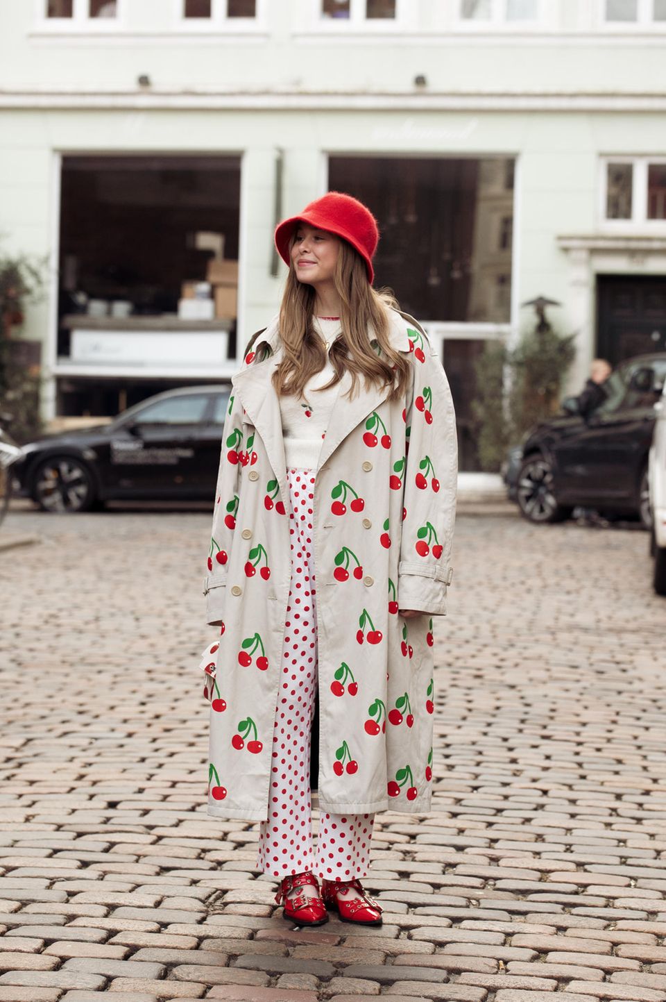 Anais van Oekel besucht die Modenschauen in einem süßen Look aus Kirschen. Ihr Trenchcoat sowie ihr Pullover werden von der Frucht geziert und ihre Hose ist komplett rot gepunktet. Extra stylisch sind ihre roten Lederschuhe mit Silberschnallen.
