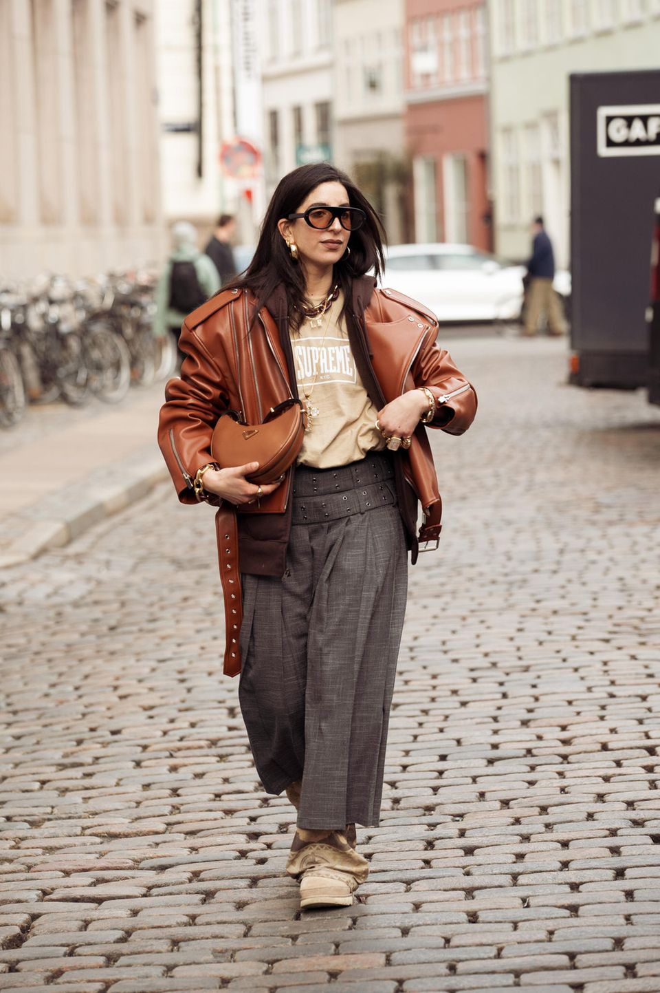 Wie man stylisch Brauntöne kombiniert, zeigt Selma Kaci mit diesem Look in Kopenhagen. Sie trägt eine kamelfarbene Lederjacke, ein beigefarbenes T-Shirt von Supreme und kombiniert darunter eine dunkelbraune Kapuzenjacke. Ihre Sonnenbrille hat ebenfalls braun getönte Gläser und abgerundet wird der Look mit einer farblich abgestimmten Tasche.