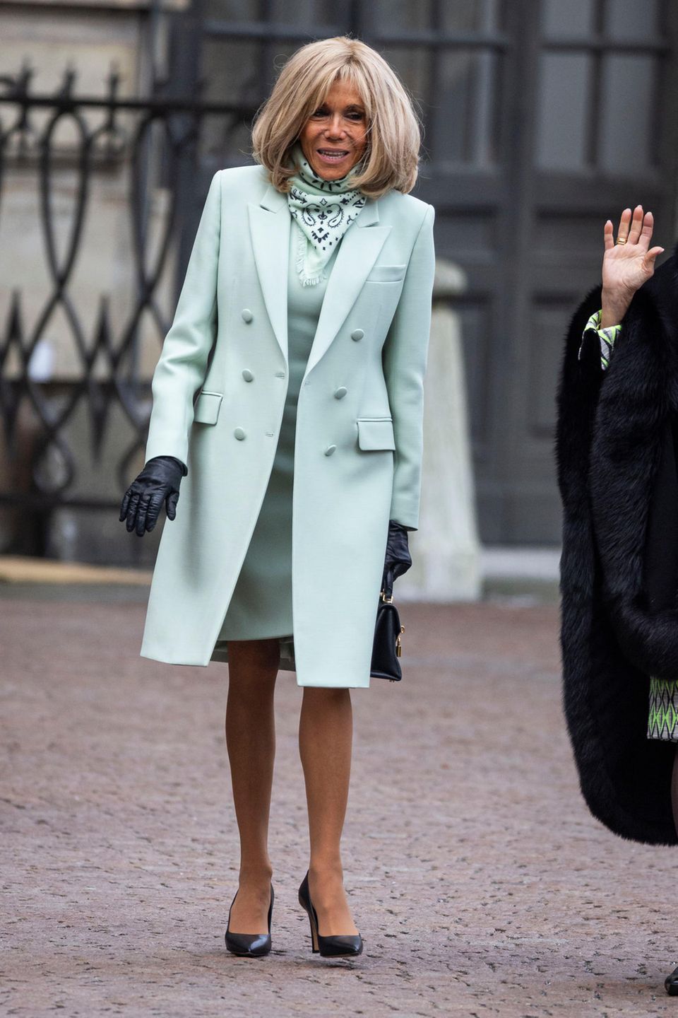 Brigitte Macron strahlt im stylischen Ton-inTon-Look auf dem Weg zum schwedischen Palast in Stockholm. Ihr Outfit besteht aus einem taillierten Mantel, einem Kleid und einem Halstuch – alles fabrlich perfekt abgestimmt. Nur ihre spitzen Pumps sowie ihre Handschuhe sorgen für einen kleinen farblichen Kontrast.