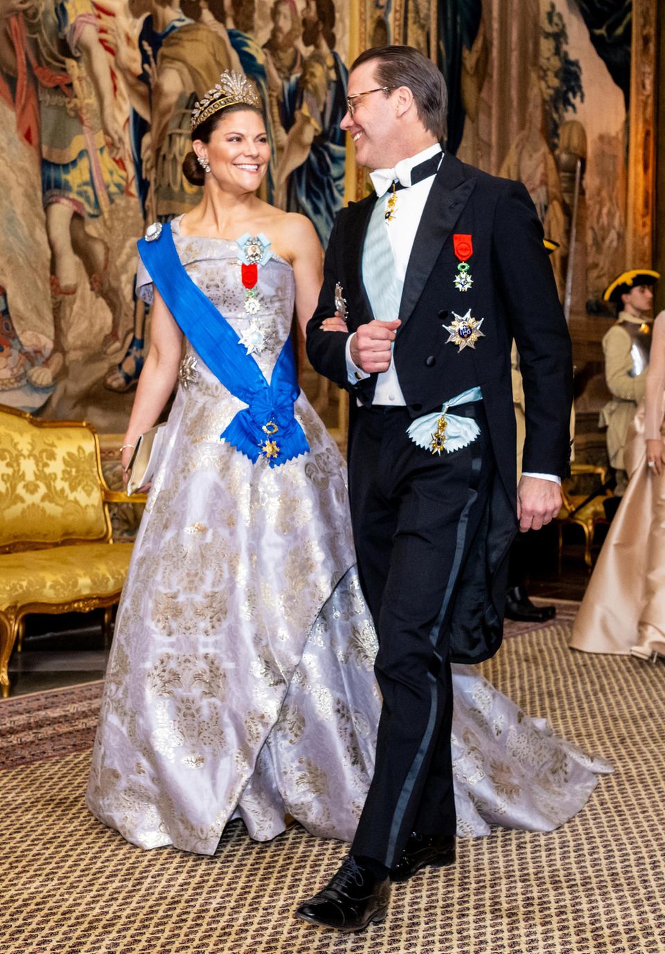 In schönster Laune schreiten auch Victoria und Daniel Richtung Festsaal. Die Kronprinzessin glänzt hierbei in einem silbergoldenen One-Shoulder-Look von H&M mit blauer Schärpe, ihr Prinz trägt ganz klassisch einen festlichen Frack.