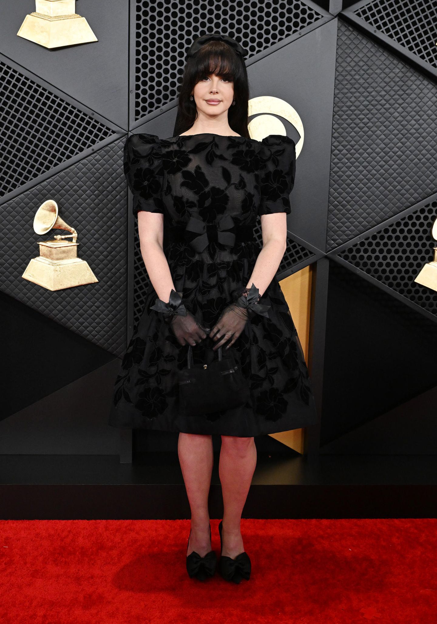 Einen dunkleren Look wählt Lana Del Rey. Sie trägt ein schwarzes Kleid mit Puffärmeln und einem Blumenmuster. Ein Hingucker sind ihre schwarzen, transparenten Handschuhe und ihr Haarreifen.