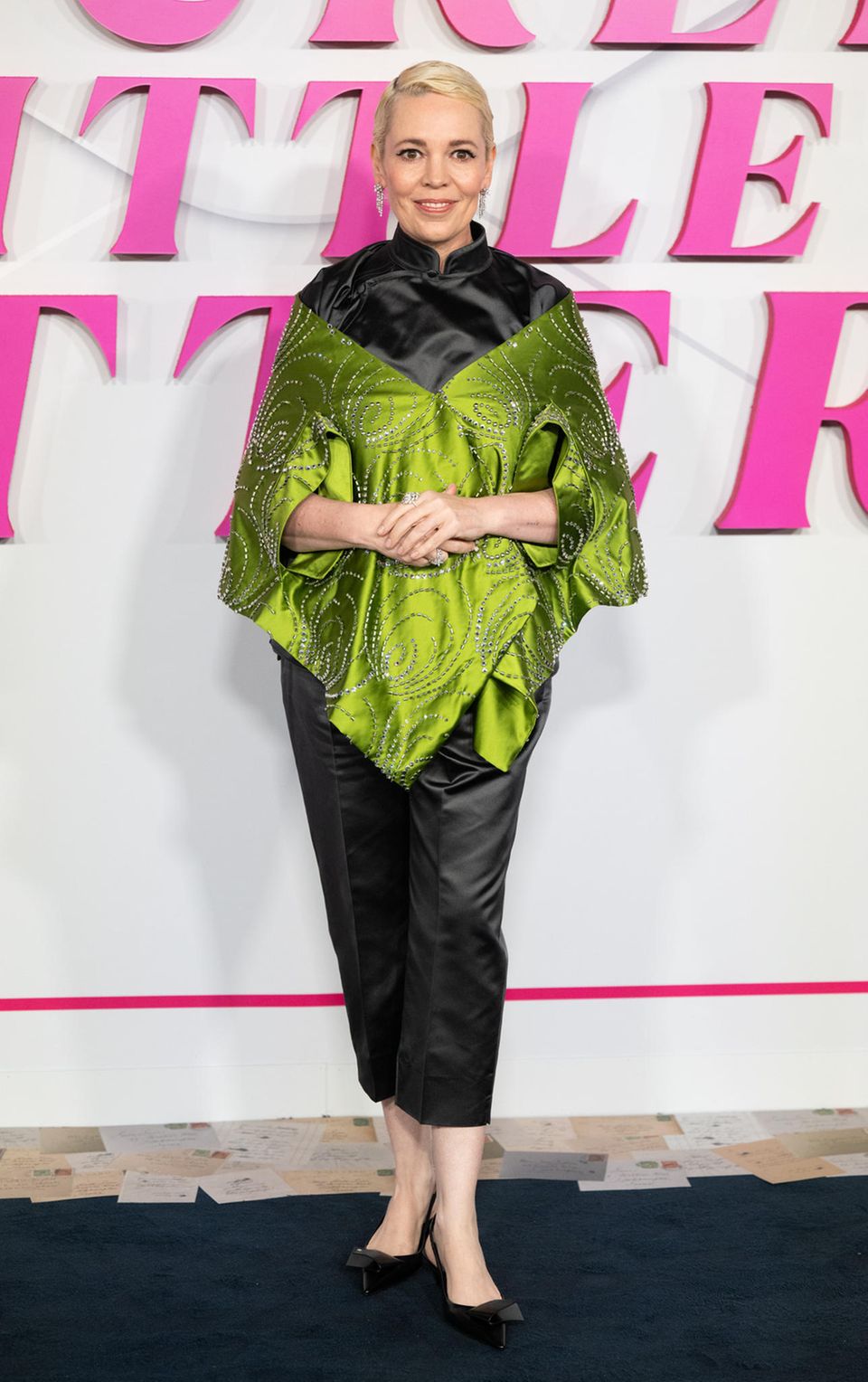 In einem extravaganten Look besucht Olivia Colman die "Wicked Little Letters"-Premiere in London. Ihr ponchoartiges Oberteil glänzt in einem Kontrast aus Grün und Schwarz mit einigen Glitzerdetails. Dazu kombiniert sie eine Chinohose aus Satin und Pumps.
