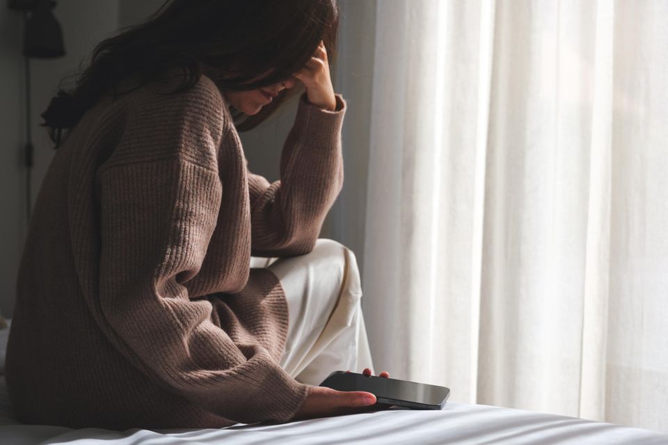 Unglücklich verliebt: Unglückliche Frau sitzt auf dem Bett und start traurig auf ihr Handy