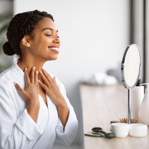 Make-up richtig auftragen: Junge Frau guckt lächelnd in den Spiegel und berührt ihren Hals