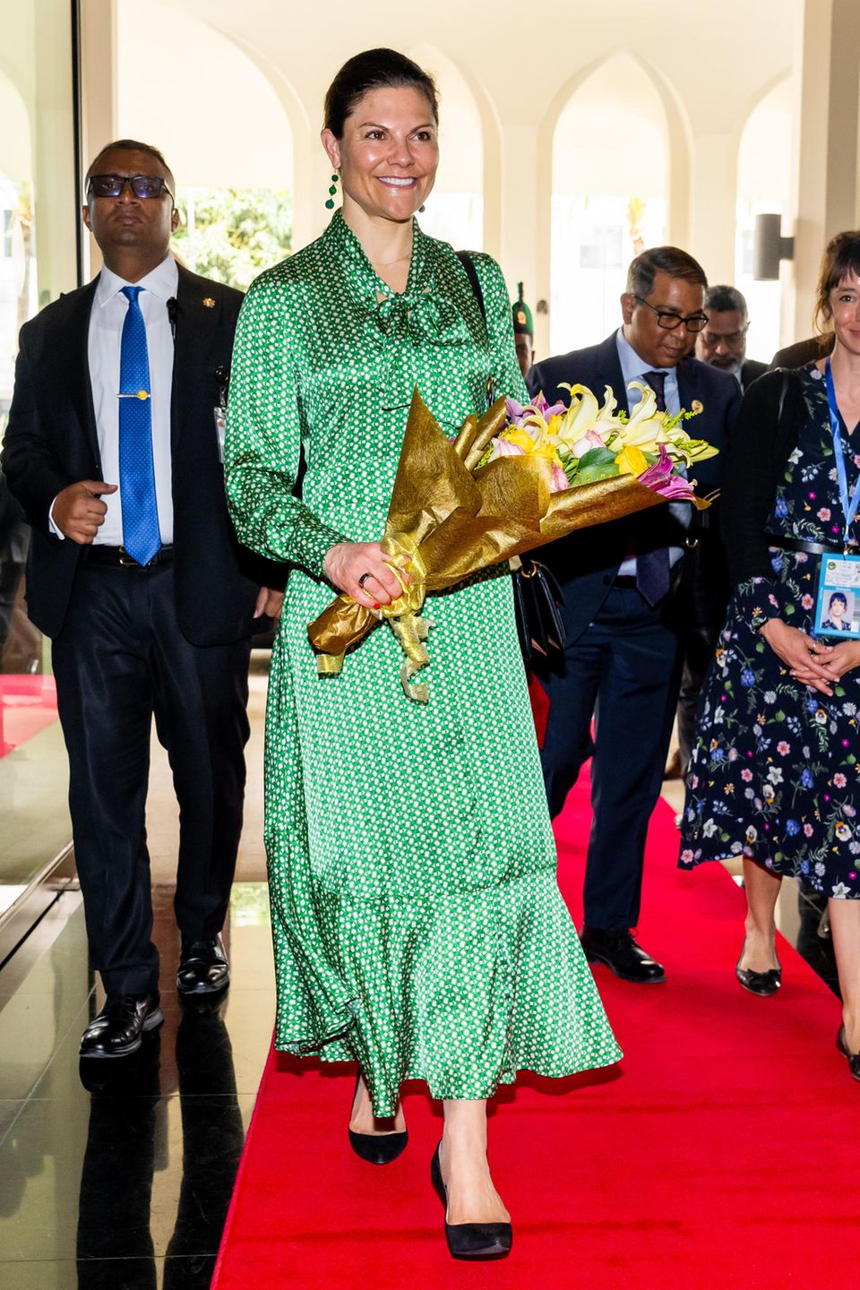 In einem verspielten, aber eleganten Maxidress von Dea Kudibal stattet Kronprinzessin Victoria von Schweden Bangladesch einen viertägigen Besuch ab. Sie fungiert bei diesem Besuch in ihrer Funktion als Sonderbotschafterin des Entwicklungsprogramms der Vereinten Nationen (UNDP). Zu ihrem grünen Kleid kombiniert sie passende Ohrringe mit grünen Steinen und schwarze Pumps.