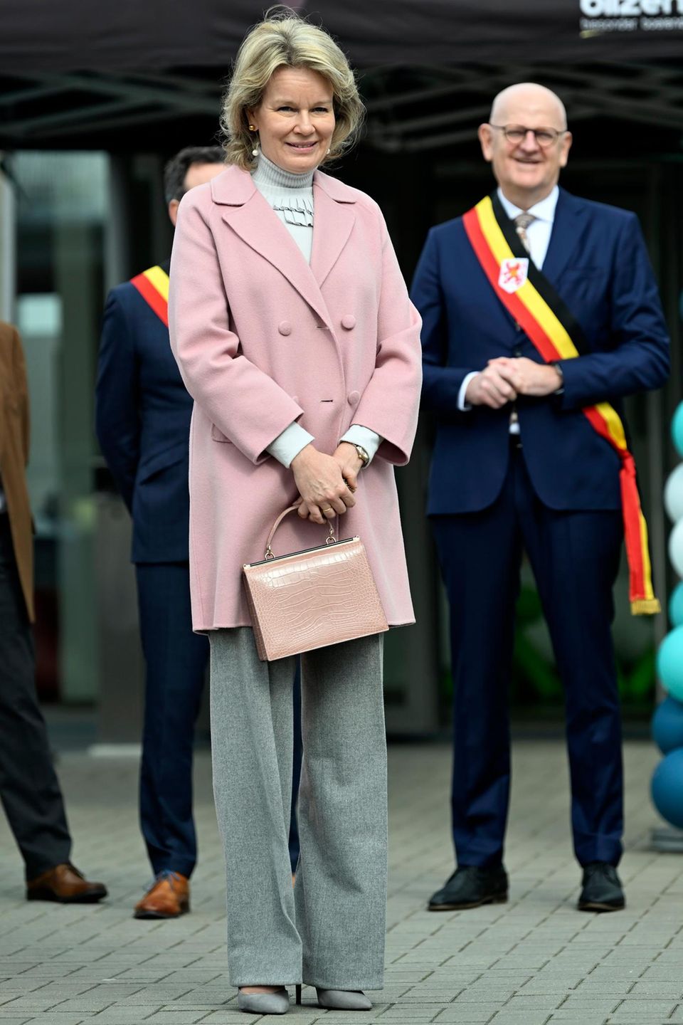 In einem stylischen, altrosafarbenen Mantel besucht Königin Mathilde von Belgien die Kinder- und Jugendabteilung der psychiatrischen Klinik Het Medisch in Bilzen. Unter dem Mantel trägt sie ein Ensemble aus Grautönen und kombiniert dazu ebenfalls graue Pumps und Perlenohrringe.