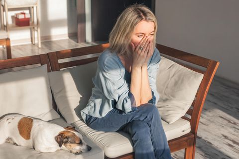 Mental Load: Erschöpfte Frau sitzt auf einem Sofa, neben ihr liegt ein Hund