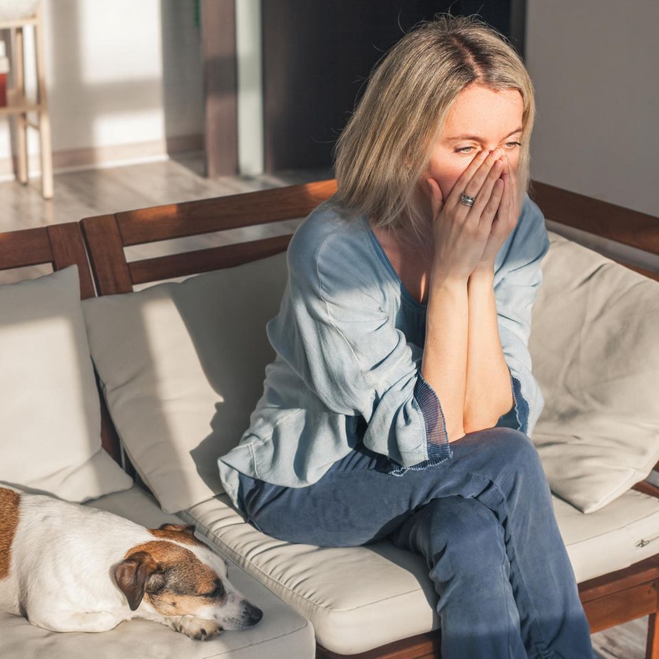 Mental Load: Erschöpfte Frau sitzt auf einem Sofa, neben ihr liegt ein Hund