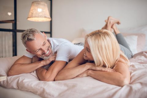 Wechseljahre und Sexualität: Ein mittelaltes Paar liegt bäuchlings auf dem Bett und lächelt sich an