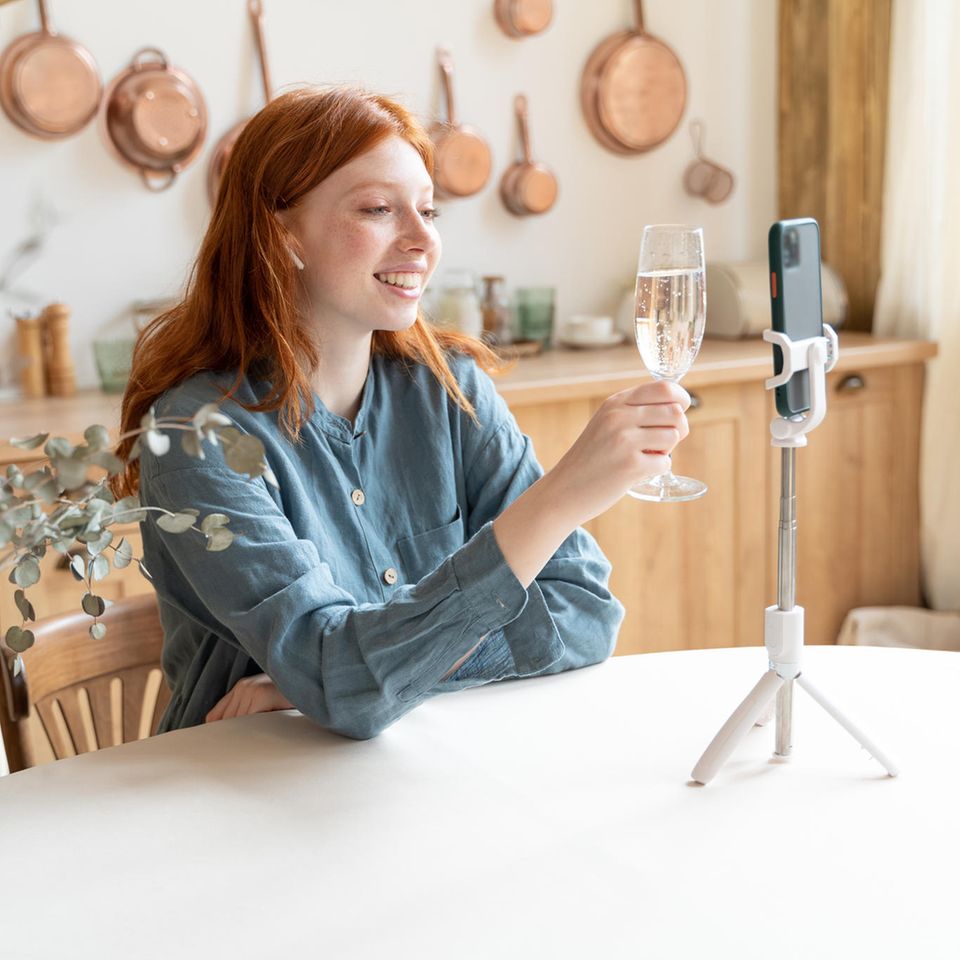 Fernbeziehung: Junge Frau sitzt am Tisch und hält ein Glas mit Sekt in der Hand, vor ihr steht ein Handy auf einem Stativ