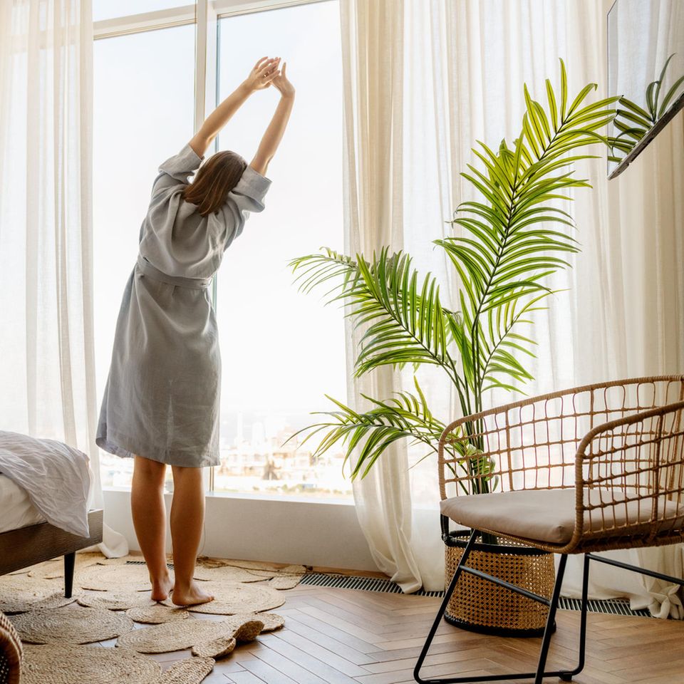 Frau streckt sich vor dem Fenster nach dem Aufstehen: 4 anstrengende Gewohnheiten, die sich wirklich lohnen