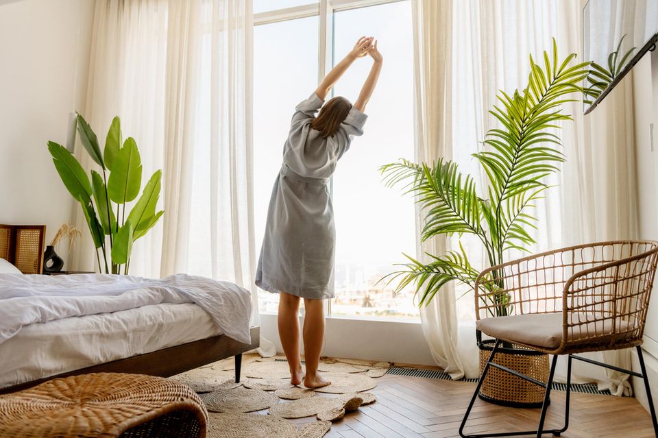 Frau streckt sich vor dem Fenster nach dem Aufstehen: 4 anstrengende Gewohnheiten, die sich wirklich lohnen