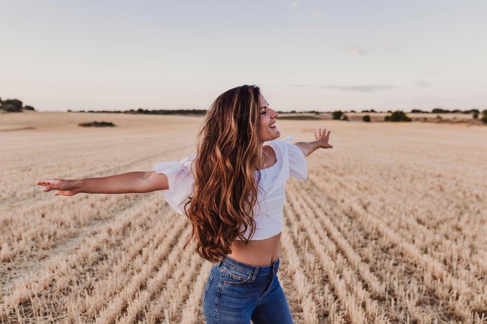 Lange Haare: Eine Frau mit langen Haaren steht auf einem Feld und breitet lächelnd die Arme aus