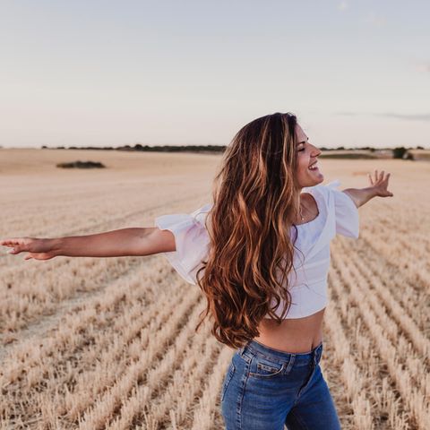 Lange Haare: Eine Frau mit langen Haaren steht auf einem Feld und breitet lächelnd die Arme aus