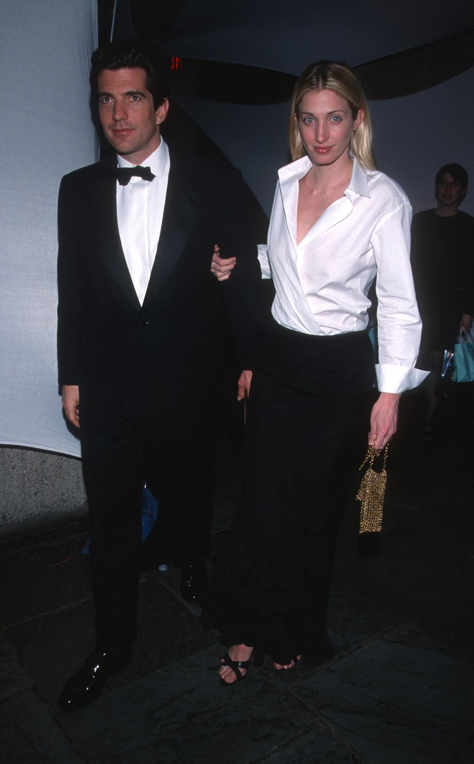 Carolyn Bessette-Kennedy is considered a style icon of the 90s. Alongside her husband, John F. Kennedy Jr., she is wearing a white shirt-blouse with a low-cut neckline tucked into her trousers. She pairs it with black trousers.